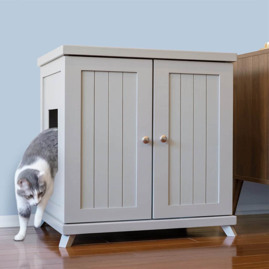 A gray and white cat steps out of a rectangular opening in a light gray litter box cabinet with two doors on a wood floor.
