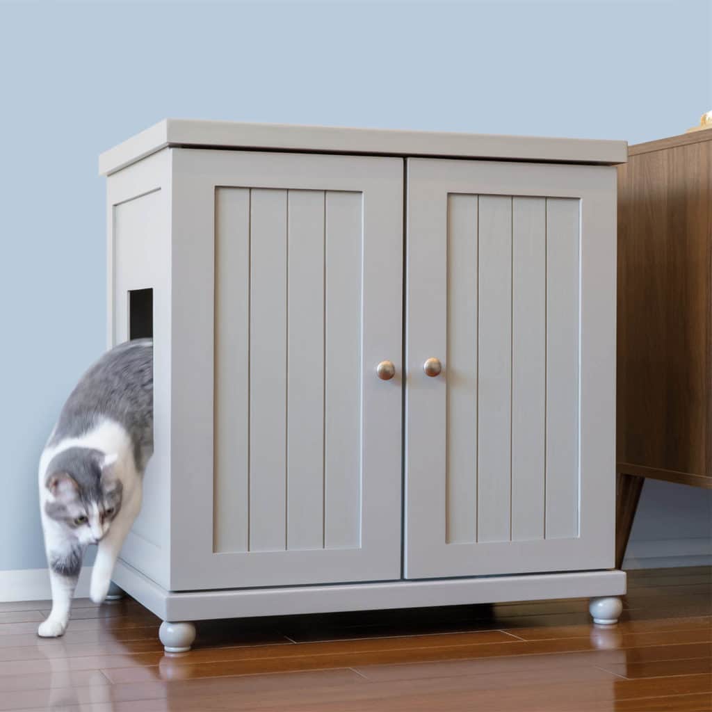 Gray and white cat exits side opening of light gray cabinet with two doors, on a wooden floor.