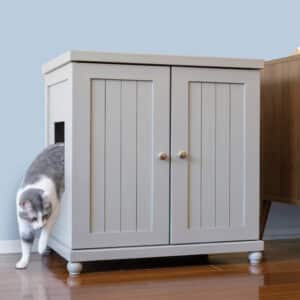 Gray and white cat exits side opening of light gray cabinet with two doors, on a wooden floor.