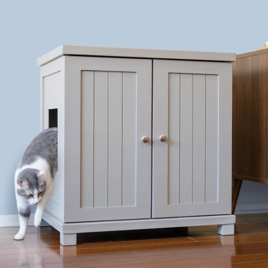 Gray and white cat steps out of a side-entrance litter box cabinet on wooden floor beside another piece of furniture.
