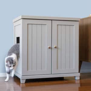 Gray and white cat exits a litter box cabinet through a side opening on a wooden floor next to other furniture.