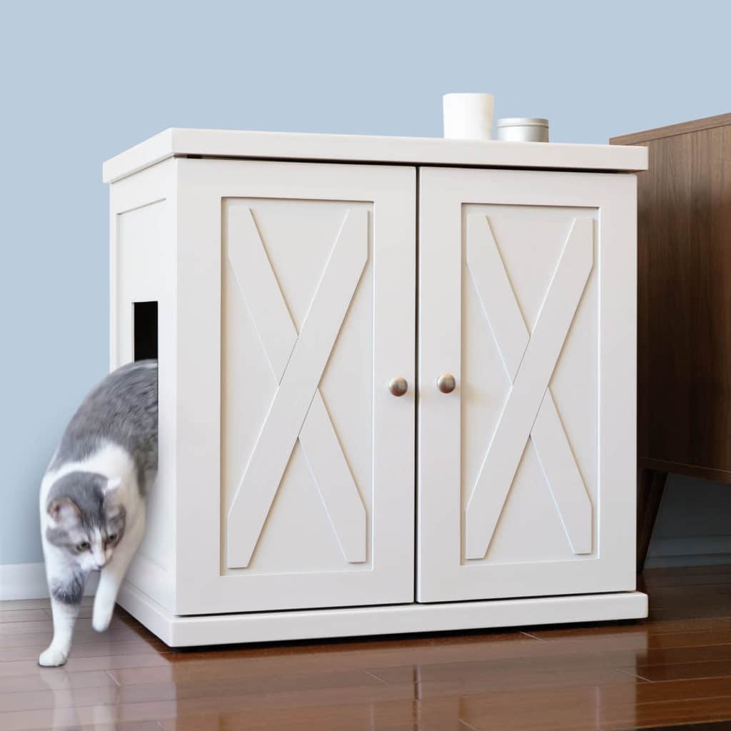A gray and white cat leaves a white litter box cabinet with double X-panel doors on a wooden floor.
