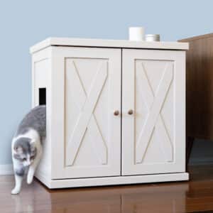 A gray and white cat leaves a white litter box cabinet with double X-panel doors on a wooden floor.