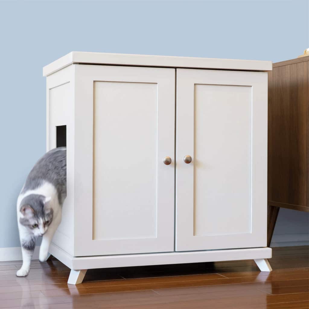 A gray and white cat exits a side opening of a white cabinet-style litter box on a wooden floor.