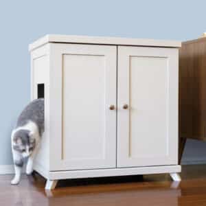 A gray and white cat exits a side opening of a white cabinet-style litter box on a wooden floor.