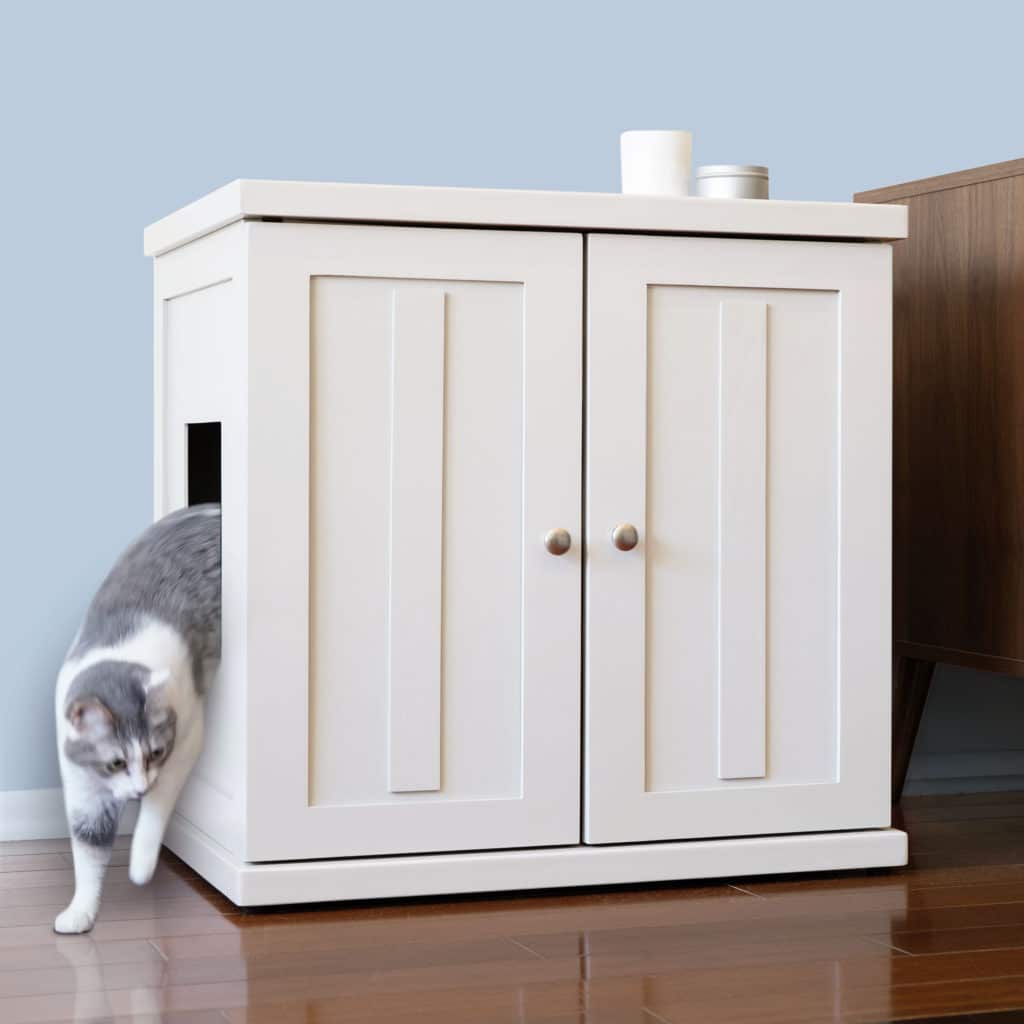 A gray and white cat leaves a white litter box cabinet with a side opening on a wooden floor beside brown furniture.