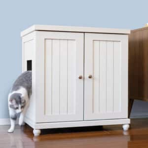 A gray and white cat steps out of a white litter box cabinet with double doors on a wooden floor.