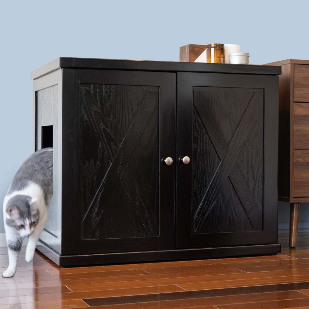 A gray and white cat leaves a litter box cabinet with double doors, on a wooden floor beside another piece of furniture.