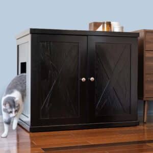 A gray and white cat leaves a litter box cabinet with double doors, on a wooden floor beside another piece of furniture.