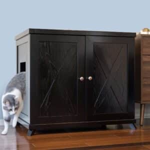 Gray and white cat exits a black litter box cabinet with double doors and side entrance on a hardwood floor near furniture.
