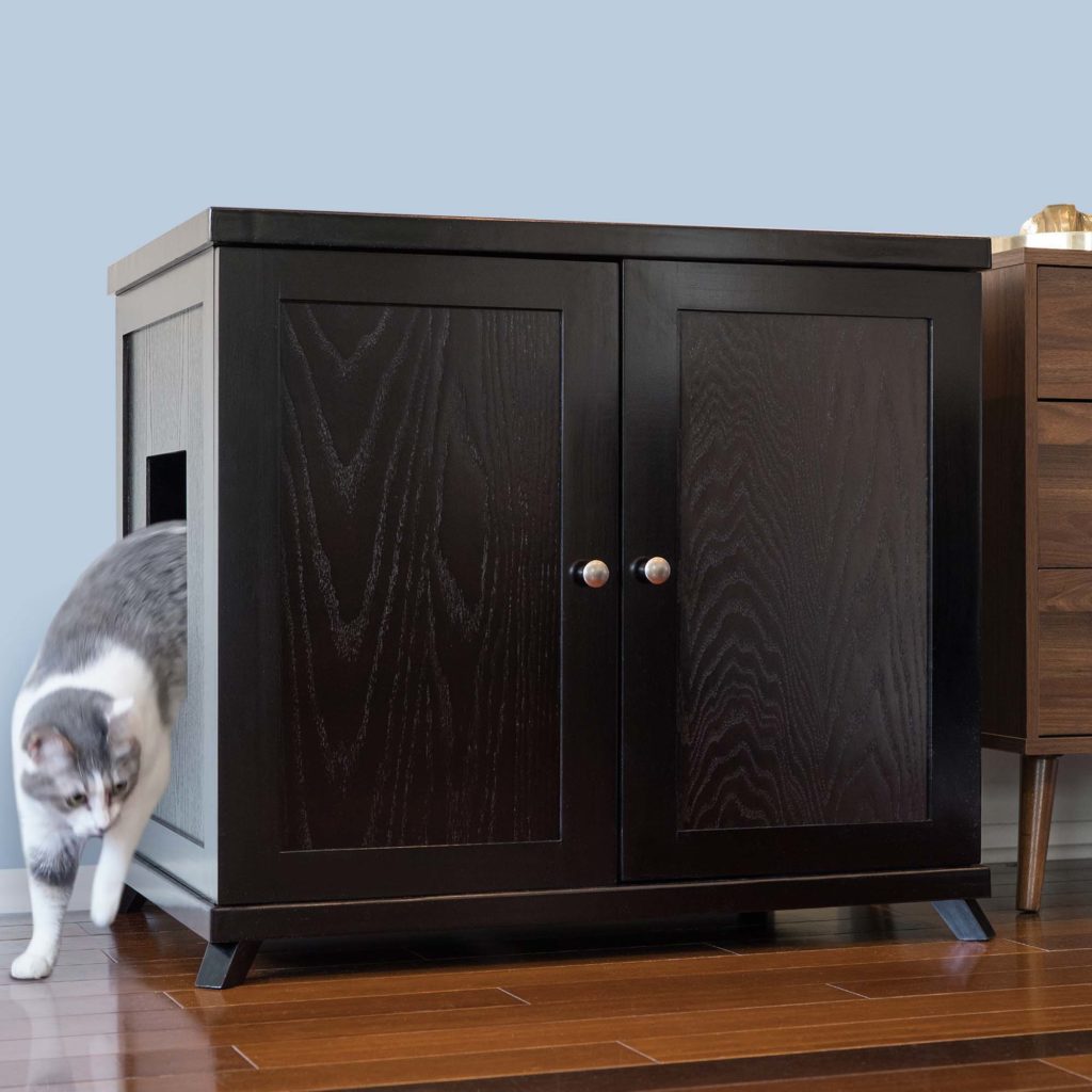 A gray and white cat leaving a dark wooden double-door cabinet through a side opening on a hardwood floor.