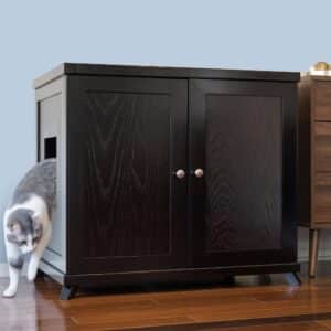 A gray and white cat leaving a dark wooden double-door cabinet through a side opening on a hardwood floor.