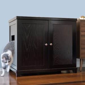 Gray and white cat exiting a black wooden litter box with side opening on hardwood floor beside another dark cabinet.
