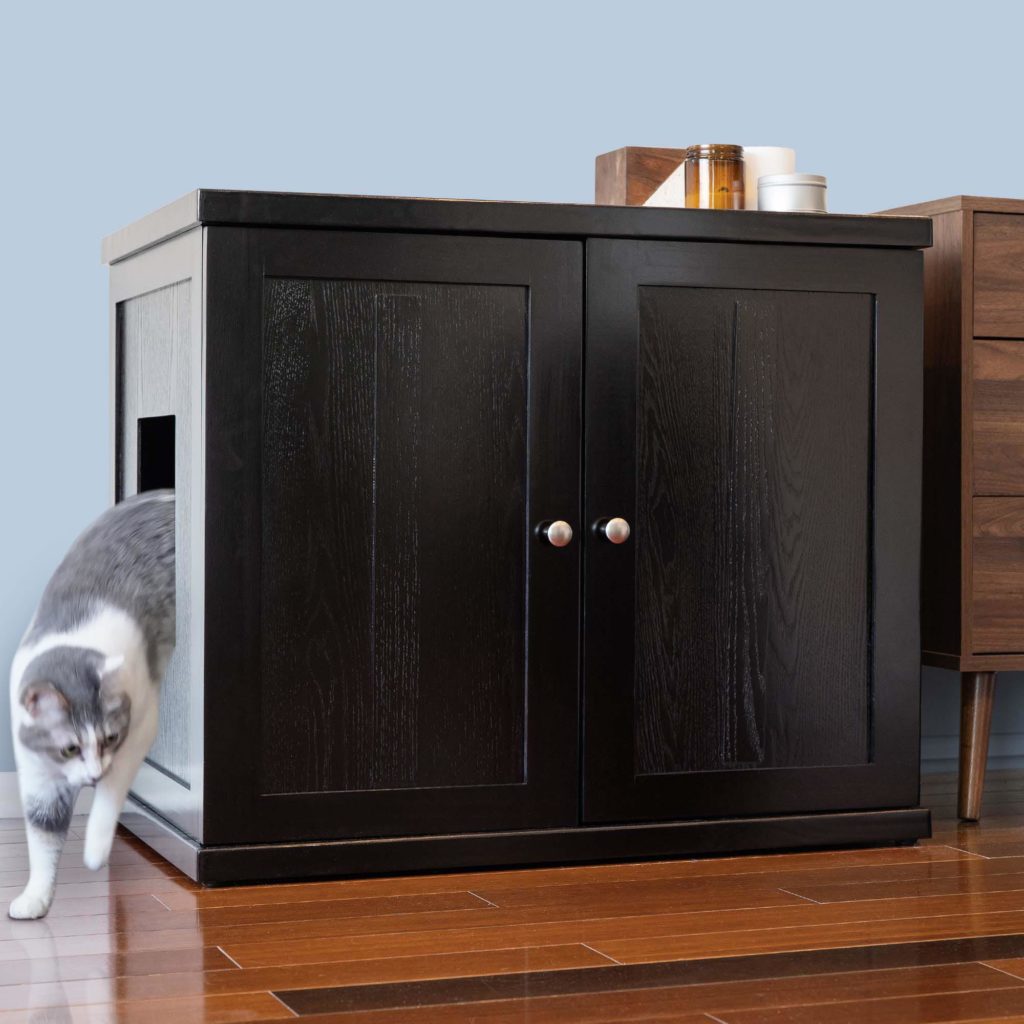 Gray and white cat stepping out of a black litter box cabinet with side opening, on hardwood floor near furniture.