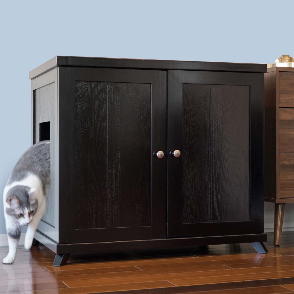 Gray and white cat steps out of a dark wood double-door cabinet on a wooden floor, hiding a litter box inside.