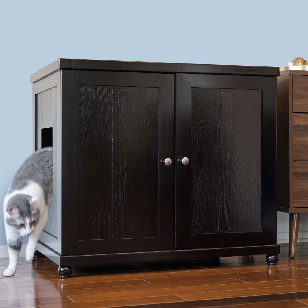 Gray and white cat steps out of a dark wood double-door litter box on wooden floor next to another furniture piece.