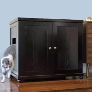 Gray and white cat steps out of a dark wood double-door litter box on wooden floor next to another furniture piece.