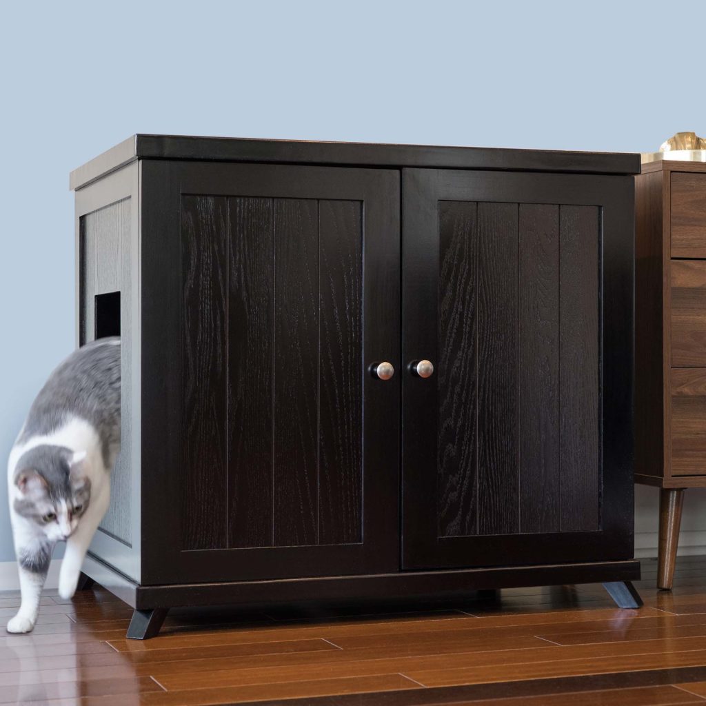 Gray and white cat exiting a dark wood litter box cabinet with side opening on hardwood floor by another wood cabinet.