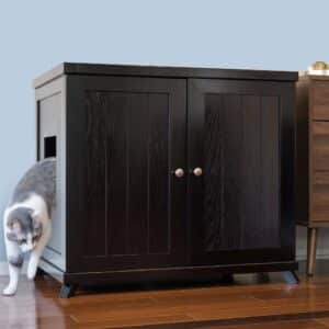 Gray and white cat exiting a dark wood litter box cabinet with side opening on hardwood floor by another wood cabinet.