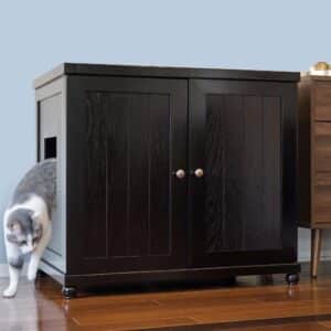 A gray and white cat steps out of a round litter box cabinet on a hardwood floor next to another cabinet.
