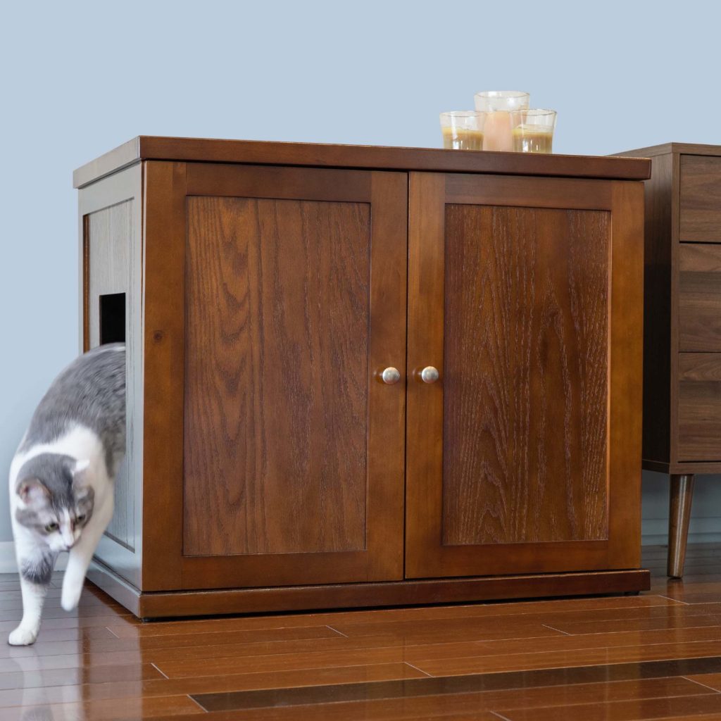 Gray and white cat walks out of a wooden cabinet-style litter box with closed doors and candles on top, on wooden floor.