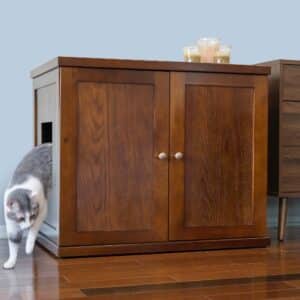 Gray and white cat walks out of a wooden cabinet-style litter box with closed doors and candles on top, on wooden floor.