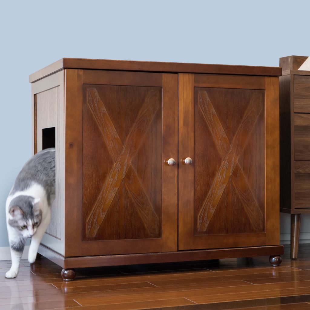 Gray and white cat walking out of a round wooden litter box cabinet on hardwood floor, against a light blue wall.