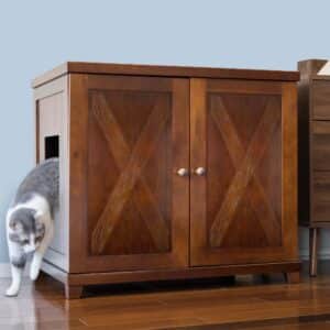 Gray and white cat exits side opening of wooden cabinet with X-patterned doors on hardwood floor, used as a litter box.