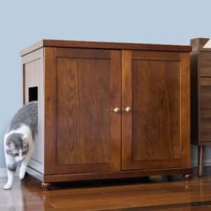 Gray and white cat exits a side-entry litter box cabinet on polished wood floor, light blue wall in background.