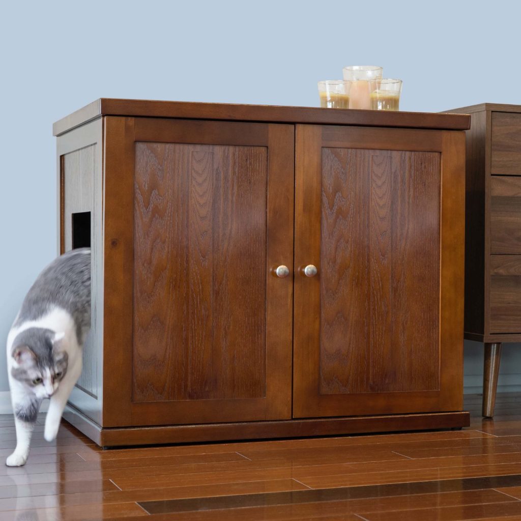 Cat stepping out of a side opening in a double-door litter box cabinet with candles on top, on hardwood floor.