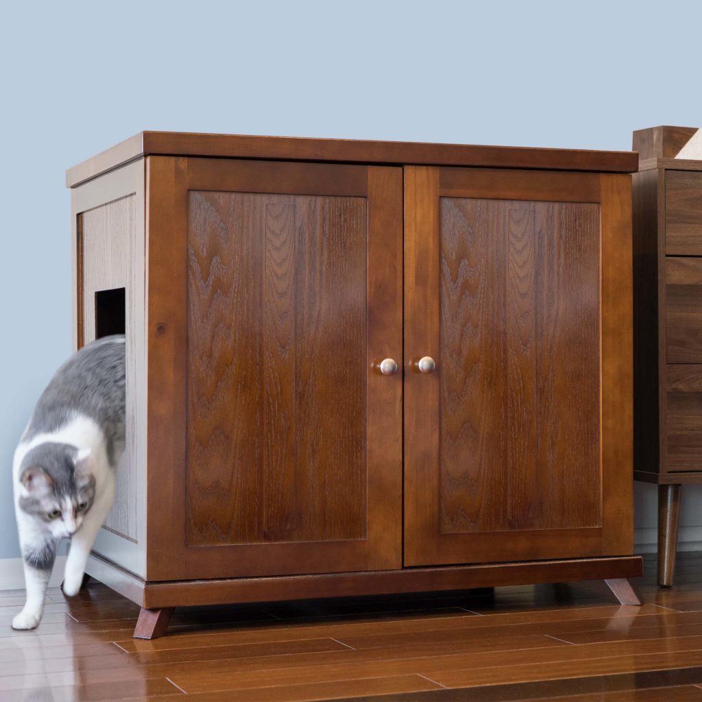 A gray and white cat leaves a wooden litter box cabinet through the side door; two front doors are closed.