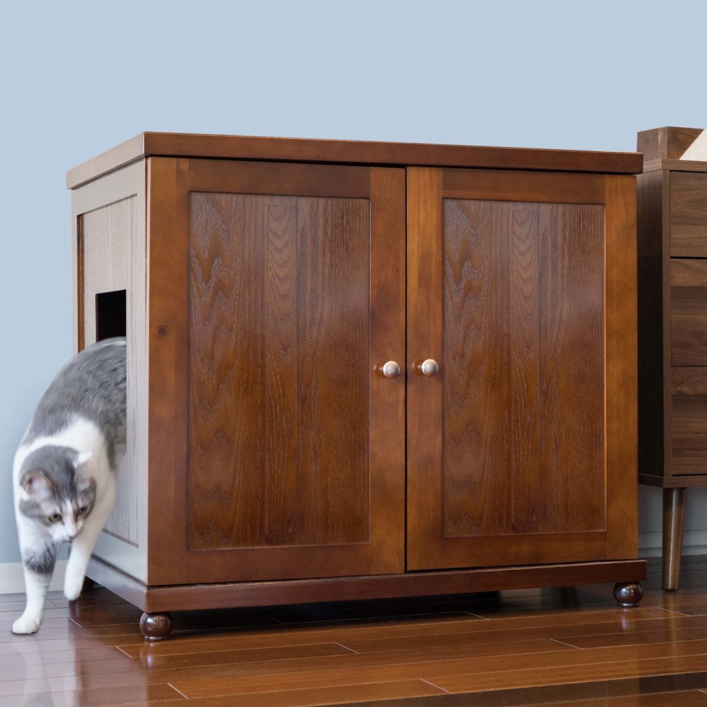 Gray and white cat exits a side opening in a round litter box cabinet with two front doors on a hardwood floor.