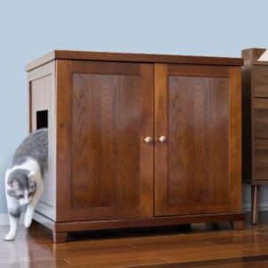 Gray and white cat exits a side opening of a wooden litter box cabinet on hardwood floor by a light blue wall.