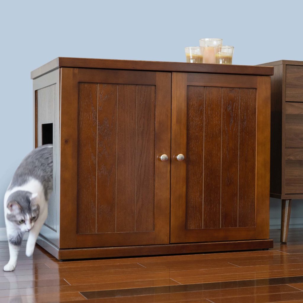 Gray and white cat exits side opening of wood cabinet litter box with candles on top, on a hardwood floor.