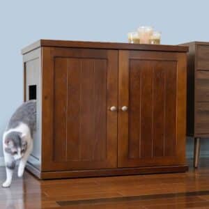 Gray and white cat exits side opening of wood cabinet litter box with candles on top, on a hardwood floor.
