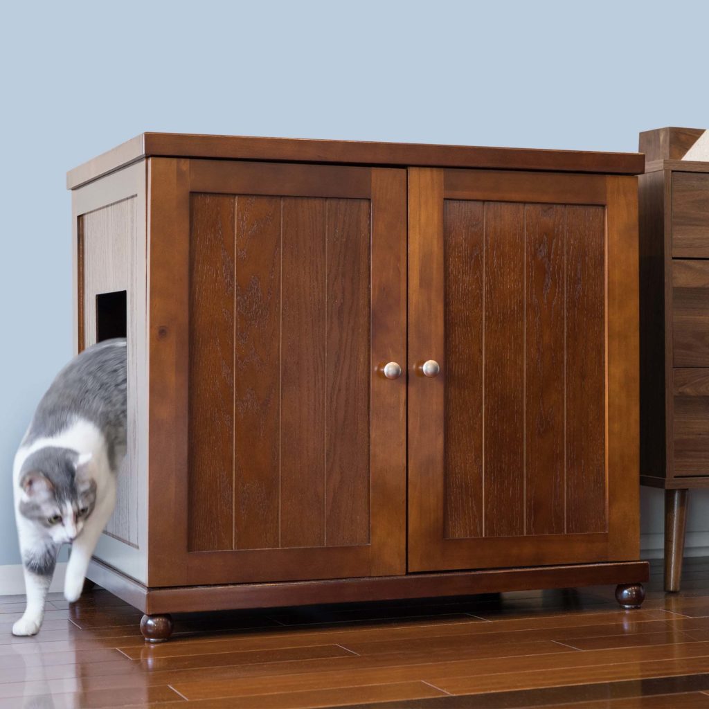 Gray and white cat exits a side compartment of a wooden litter box cabinet on a wooden floor, litter box visible inside.