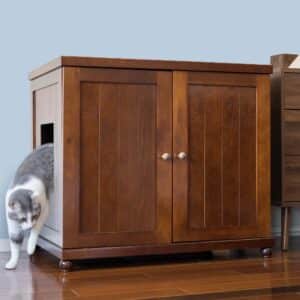 Gray and white cat exits a side compartment of a wooden litter box cabinet on a wooden floor, litter box visible inside.
