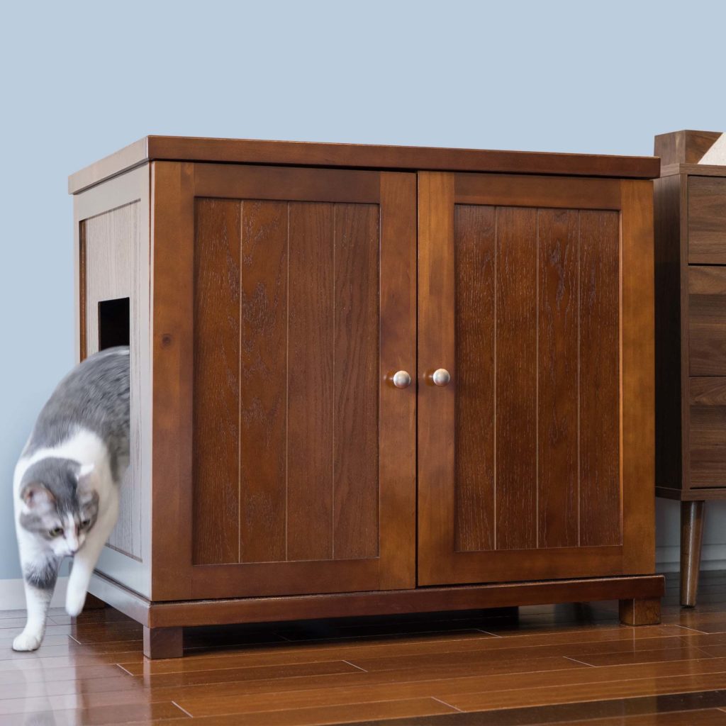 Gray and white cat exiting a side-opening litter box cabinet on hardwood floor beside a light blue wall.