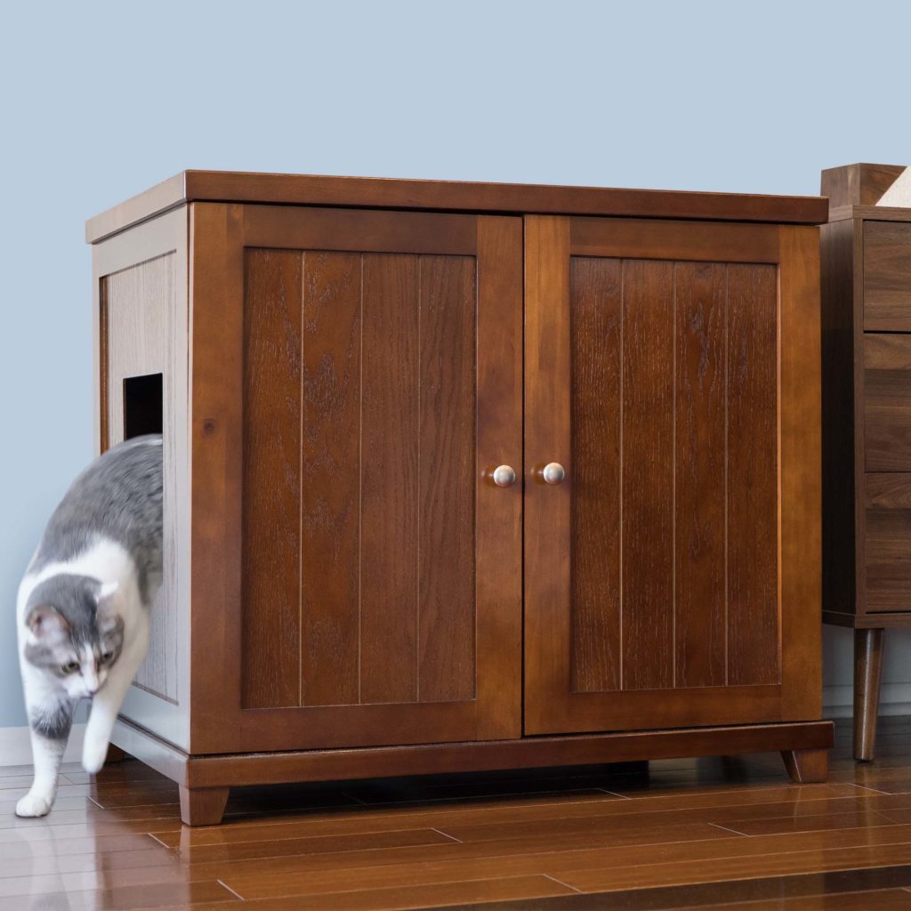Gray and white cat walking out of a wood-style double-door litter box cabinet on a hardwood floor.