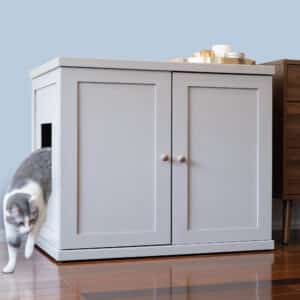 A gray and white cat steps out of a side-opening litter box cabinet on a wooden floor next to brown furniture.