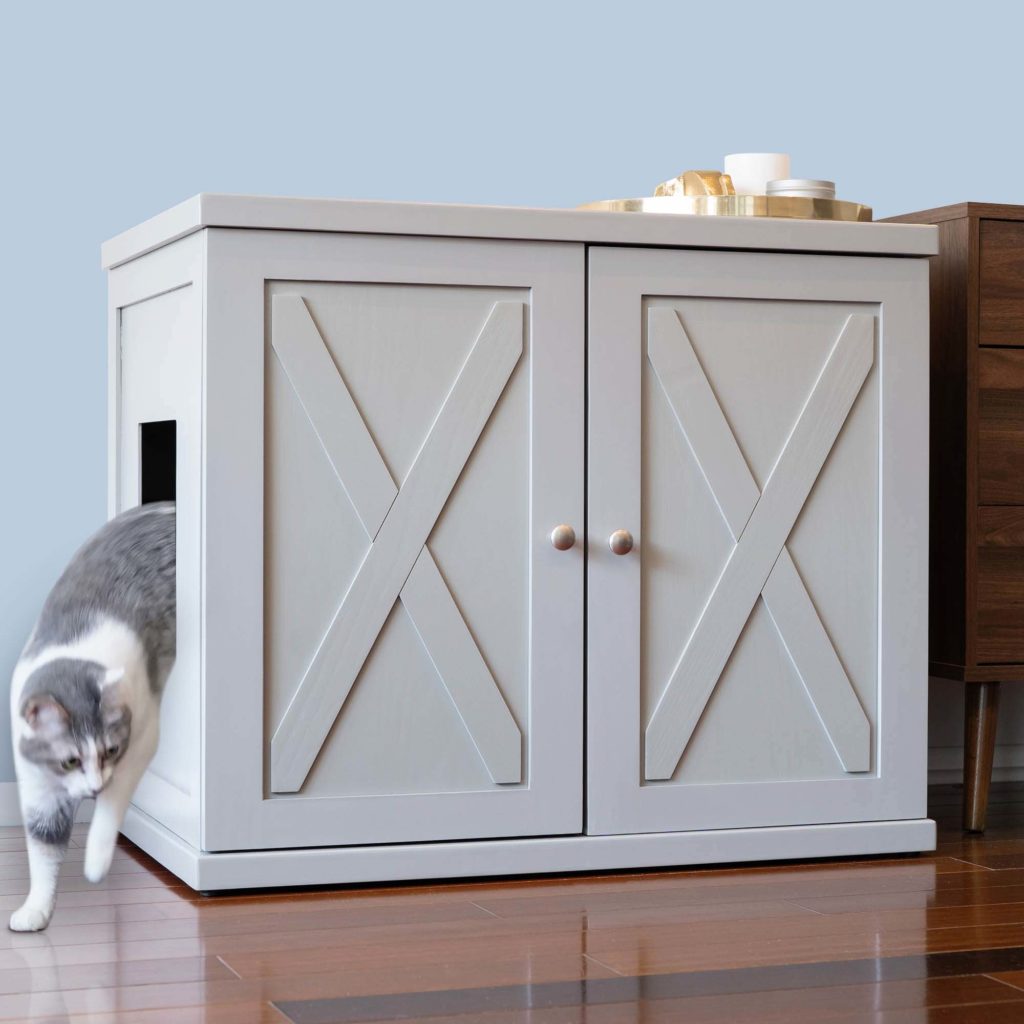 Gray and white cat exits a light gray cabinet with X-shaped doors on a wooden floor next to a wooden table.