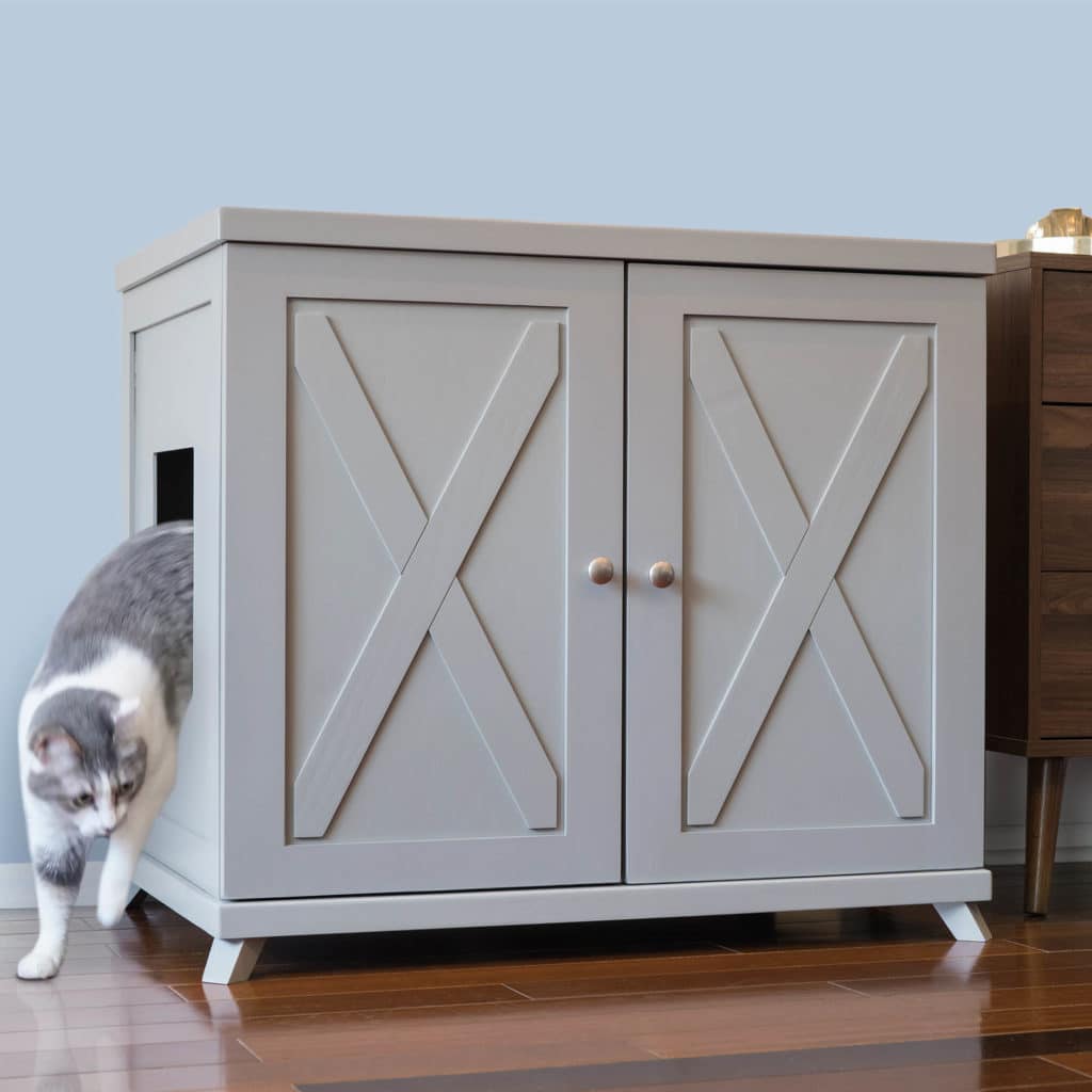 A gray and white cat steps out of a side-entry litter box cabinet on a wooden floor beside another piece of furniture.