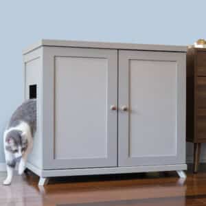 Gray and white cat steps out of a light gray two-door litter box cabinet on a wooden floor next to another cabinet.