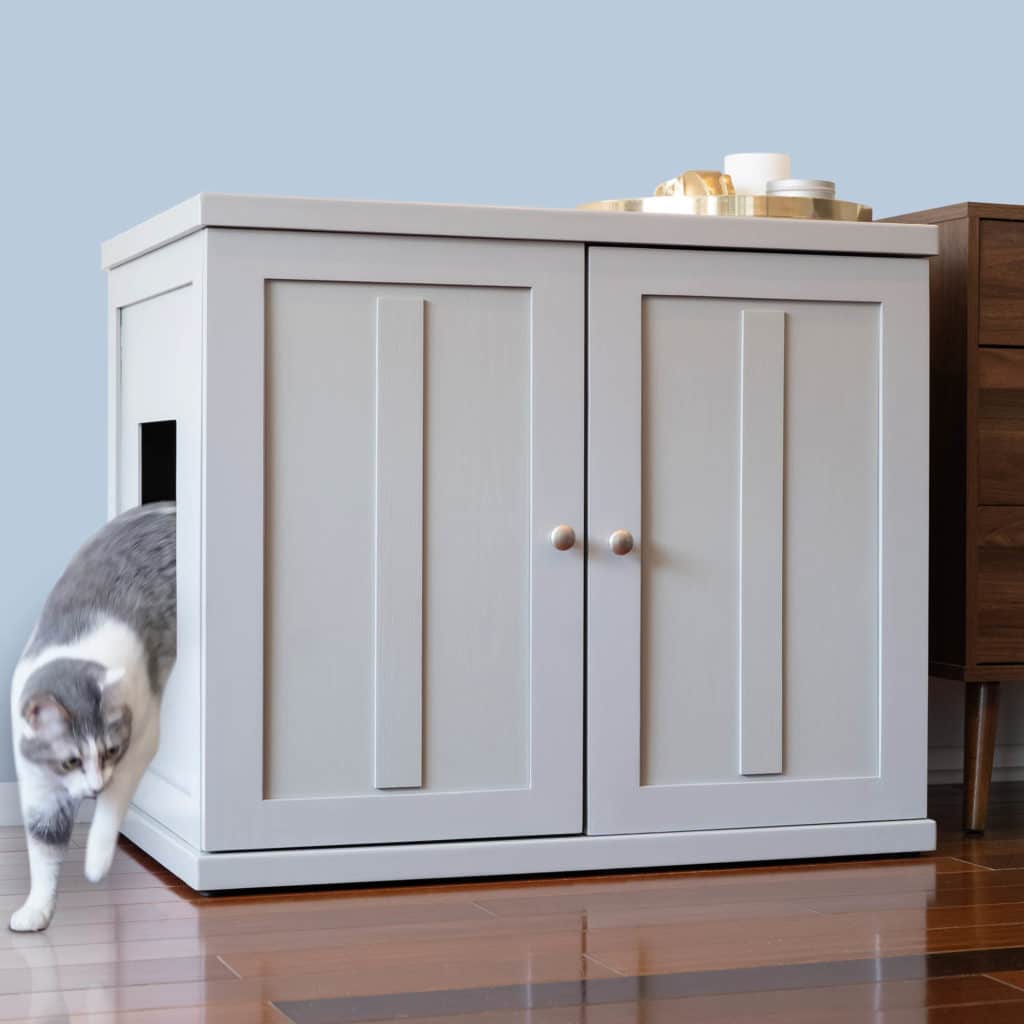 Gray and white cat exits side opening of light gray double-door litter box cabinet on a wooden floor.