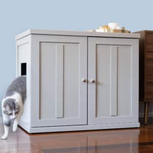 Gray and white cat exits side opening of light gray double-door litter box cabinet on a wooden floor.
