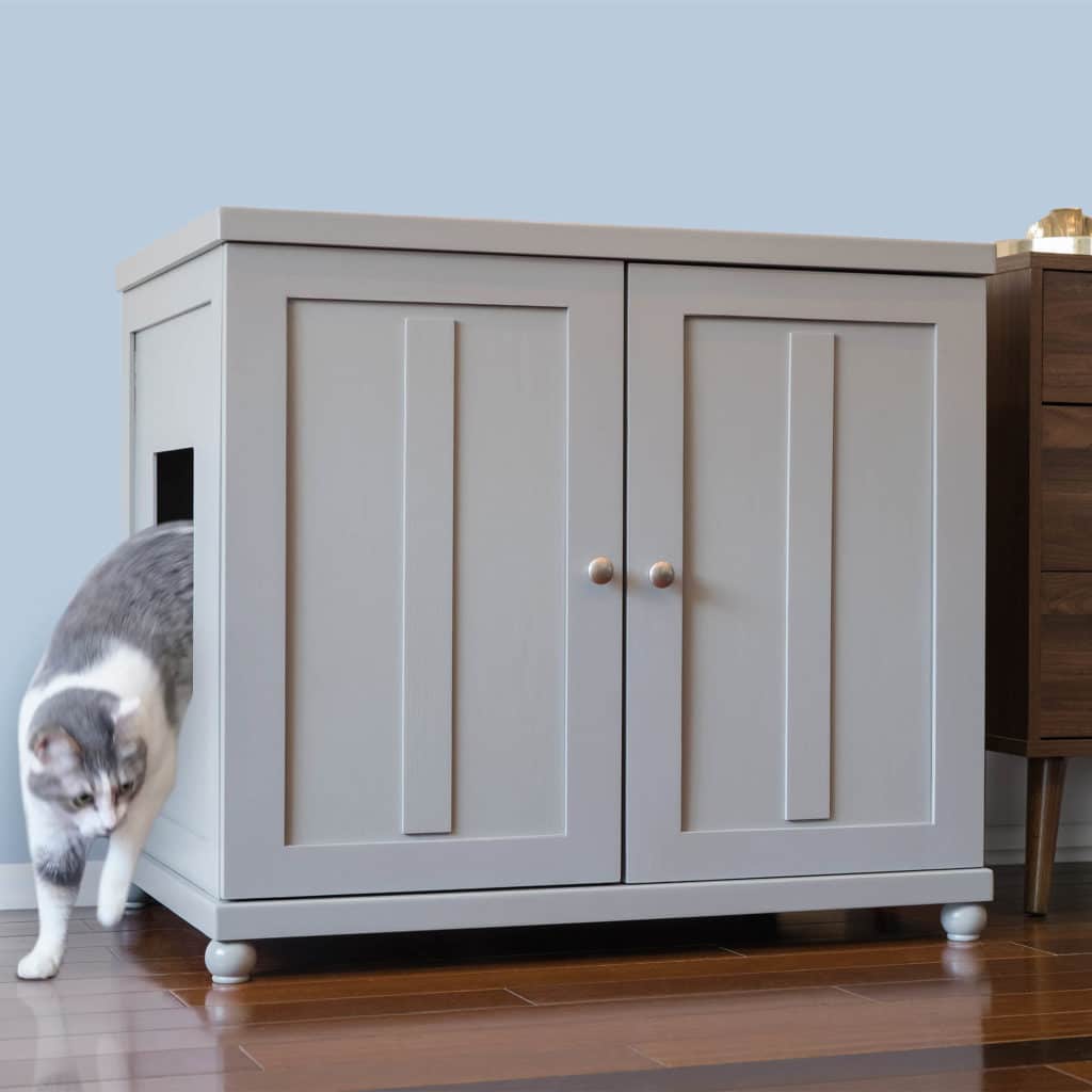 Gray and white cat leaves a light gray round litter box cabinet on wooden floor next to darker wood furniture.