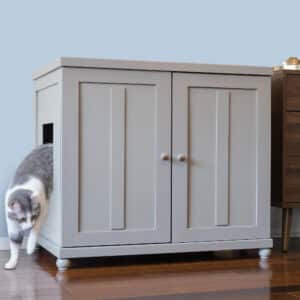 Gray and white cat leaves a light gray round litter box cabinet on wooden floor next to darker wood furniture.