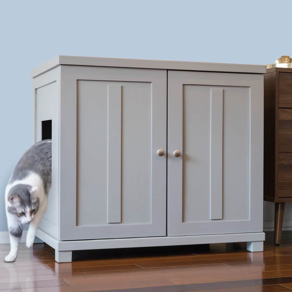 Gray and white cat walks out a side opening of a square litter box cabinet with double doors on a wooden floor.