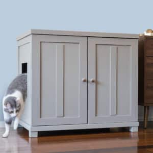 Gray and white cat walks out a side opening of a square litter box cabinet with double doors on a wooden floor.