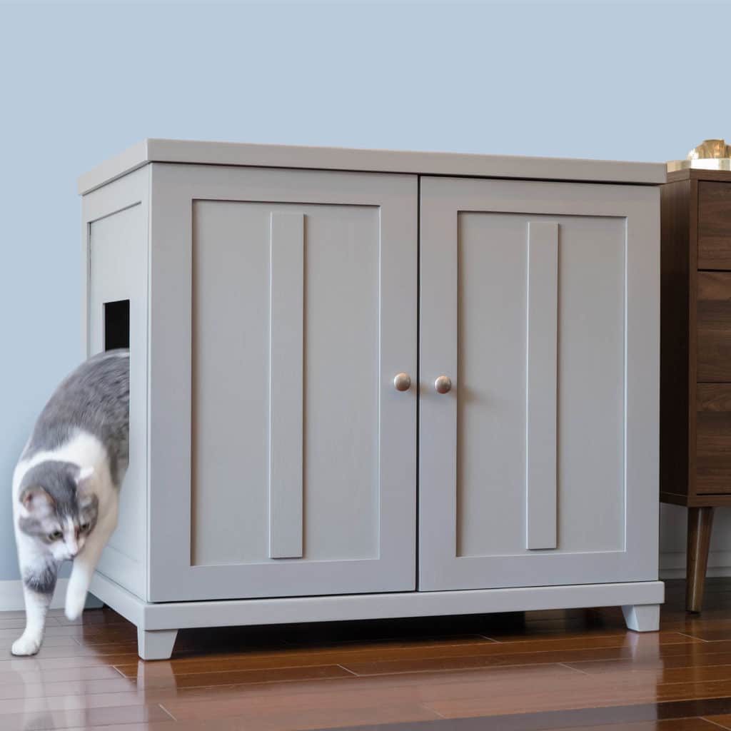 Gray and white cat steps out of a side opening in a double-door litter box cabinet on wood floor beside brown table.
